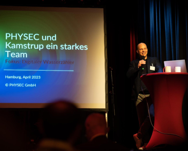 PHYSEC CFO Heiko Koepke hält eine Präsentation auf dem Kamstrup Thementag. Er steht auf der Bühne und hält ein Mikrofon in der rechten Hand. Im Hintergrund sieht man die Präsentation, die per Beamer auf eine Leinwand projiziert wird.