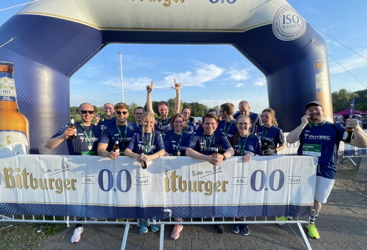 15 Mitglieder des PHYSEC Teams posieren mit ihren Medaillen bei einem Bitburger Werbetor nach Beendigung des AOK Laufs.